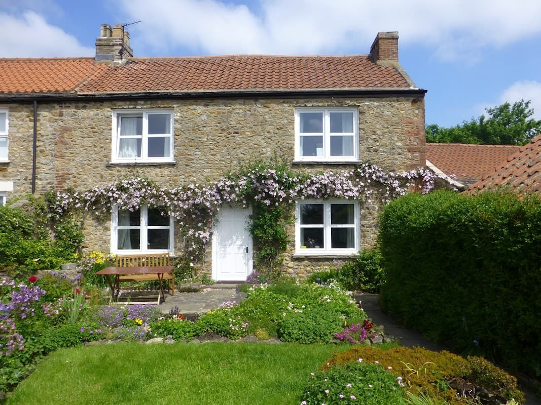 Charming stone cottage with white windows, surrounded by lush greenery and colorful flowers in Catterick, North Yorkshire.