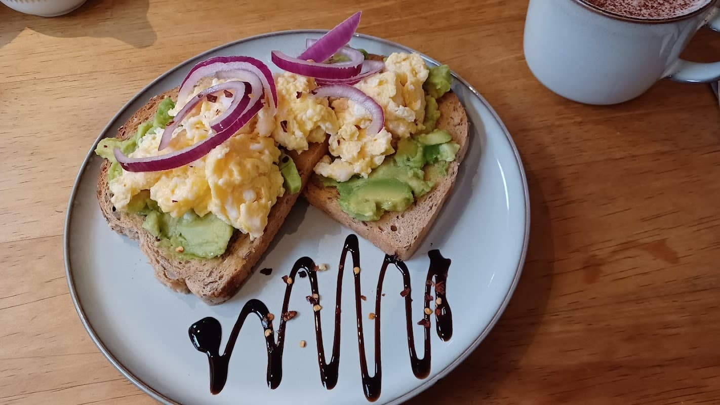 Avocado toast topped with scrambled eggs and red onion, drizzled with balsamic glaze, alongside a cup of hot chocolate.