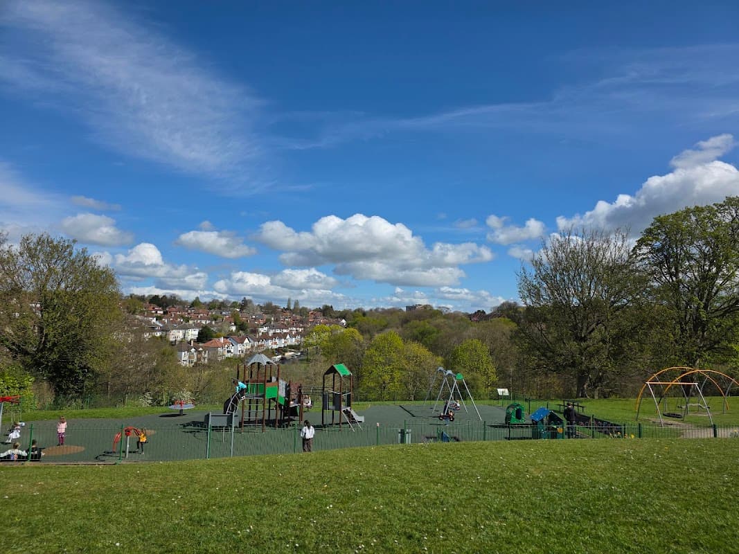 Colorful playground equipment in a park with green grass, trees, and a blue sky dotted with fluffy clouds.