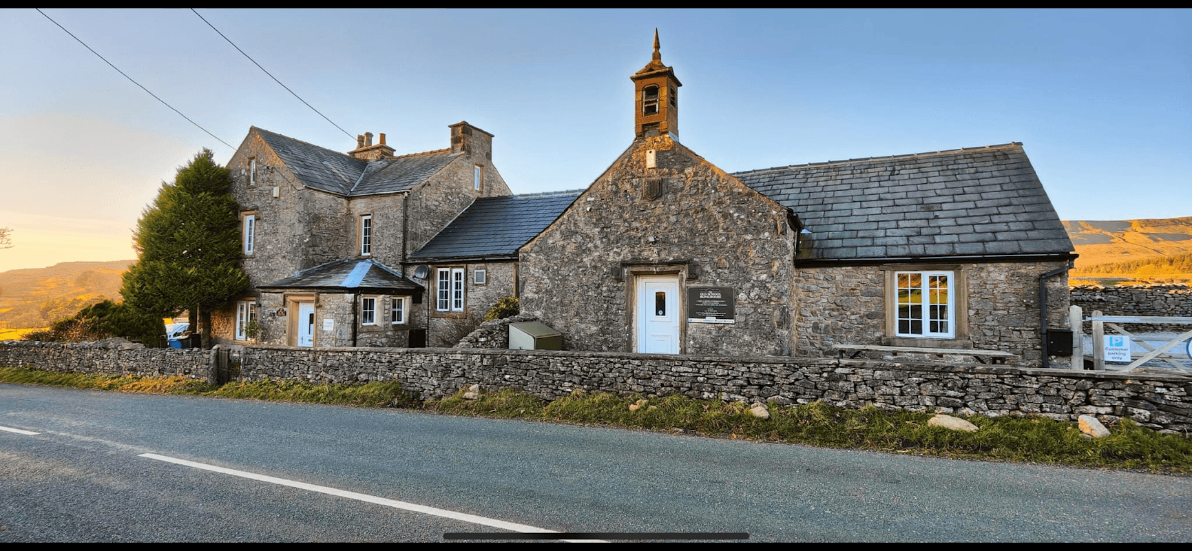 Stone building with a slate roof, bell tower, and surrounding dry stone wall, set against a scenic rural backdrop.