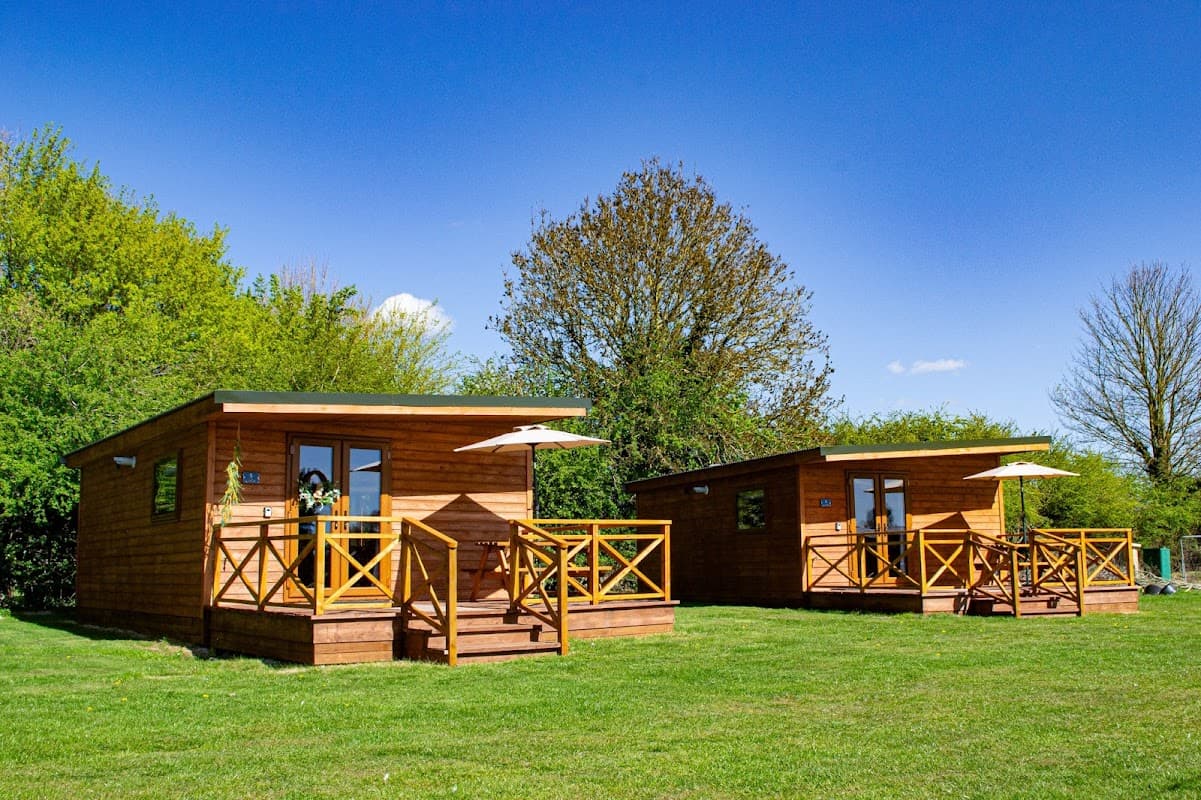 Two wooden cabins with porches on a grassy area, surrounded by trees under a clear blue sky.