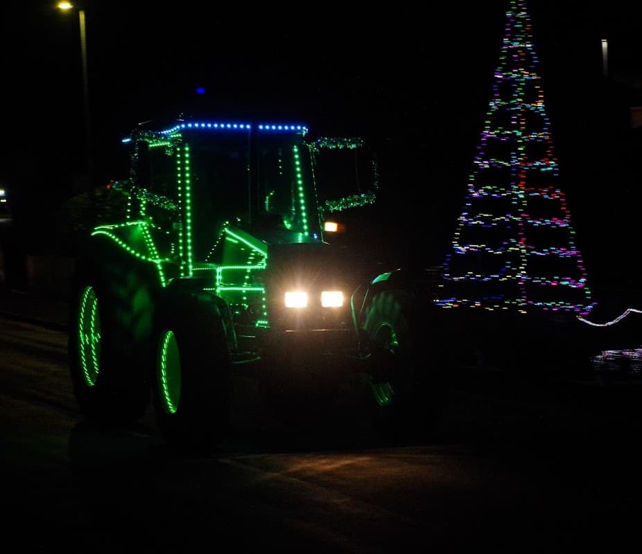 A brightly lit tractor adorned with colorful lights drives past a decorated Christmas tree at night.