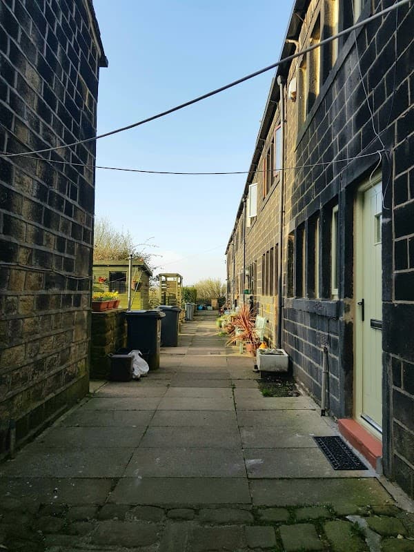 Narrow pathway between stone buildings, with planters and bins, leading to a garden area under a clear blue sky.