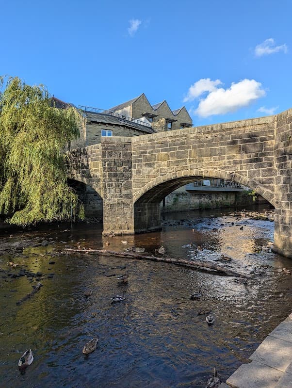 Stone bridge over a river with a weeping willow, surrounded by buildings and blue sky in Chiserley, Yorkshire.