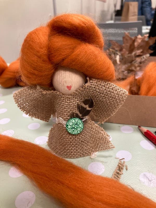 A handmade doll with orange hair, dressed in burlap, sits on a polka-dot table among crafting materials.
