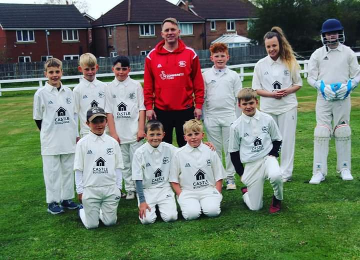 Youth cricket team in white uniforms poses on a grassy field, with a coach in a red jacket and a player in cricket gear.