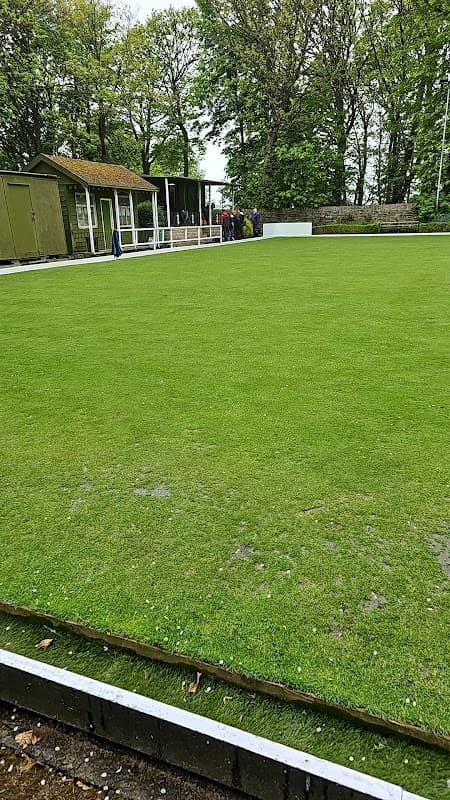 Lush green bowling green with a clubhouse in the background and people gathered under trees in Clayton West, Yorkshire.