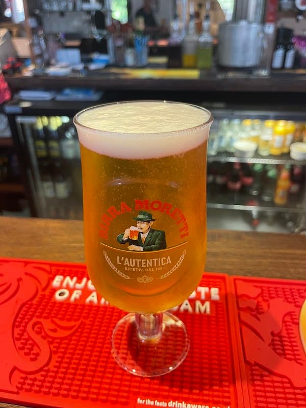 A pint of beer in a branded glass sits on a bar mat, with a blurred background of bottles and bar equipment.