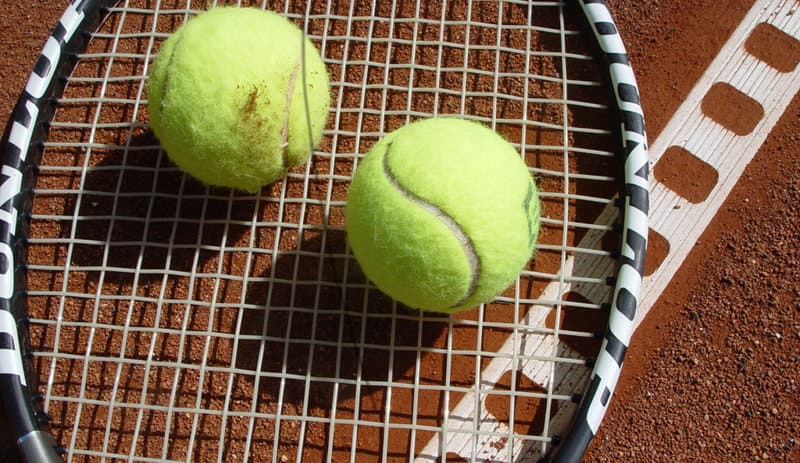 Two bright yellow tennis balls resting on a tennis racket, placed on a clay court with white lines.