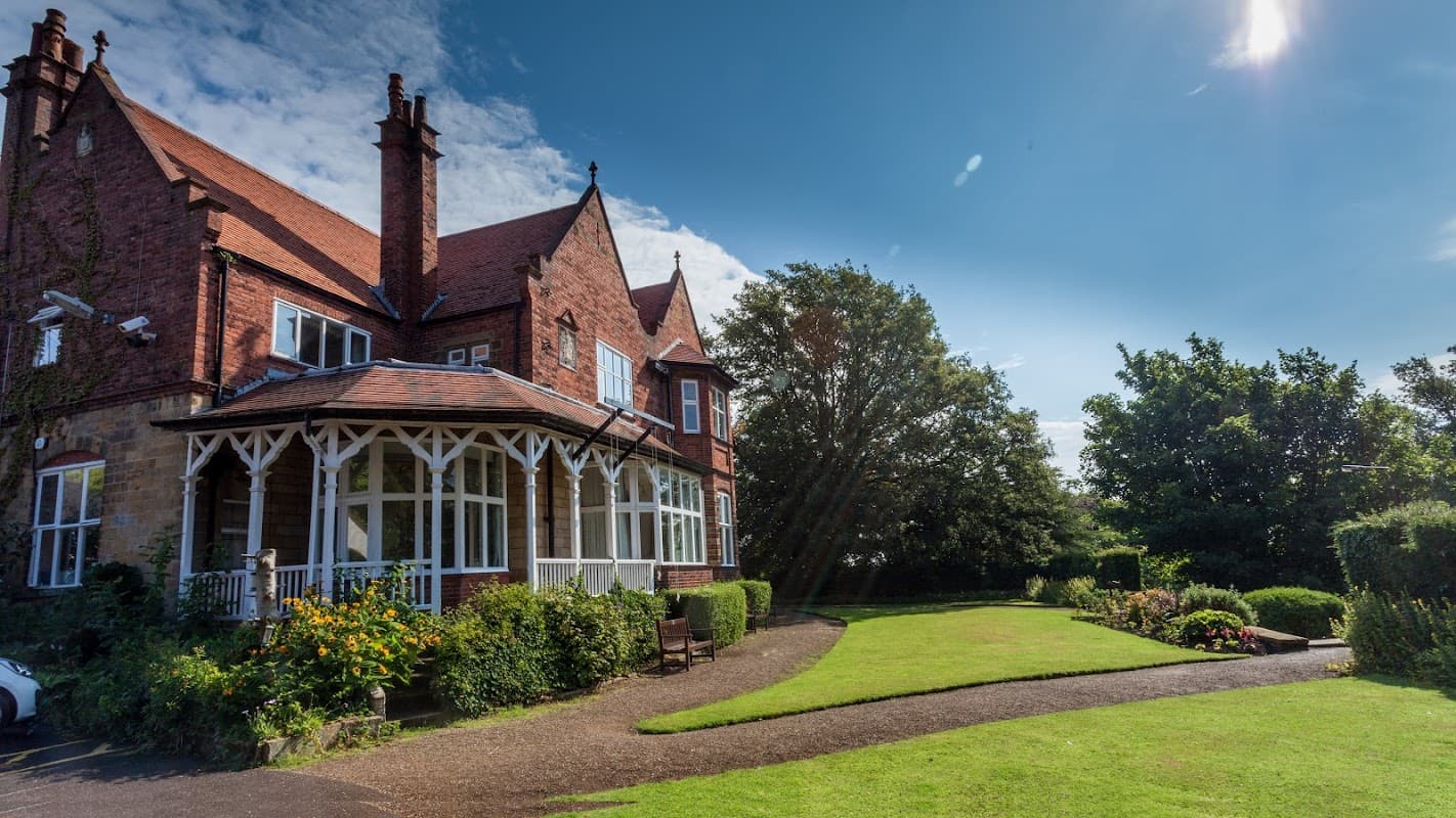 Victorian-style event venue with red brick exterior, lush green lawn, and colorful flowerbeds under a clear blue sky.