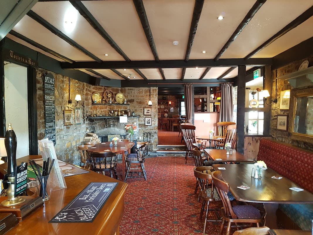 Cozy restaurant interior with wooden beams, stone walls, tables, chairs, and a fireplace, creating a warm atmosphere.