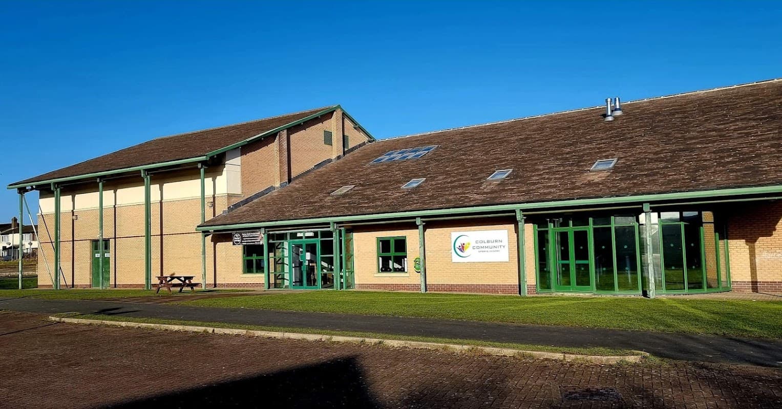 Modern sports center with large green windows, a sloped roof, and a clear blue sky in Colburn, North Yorkshire.