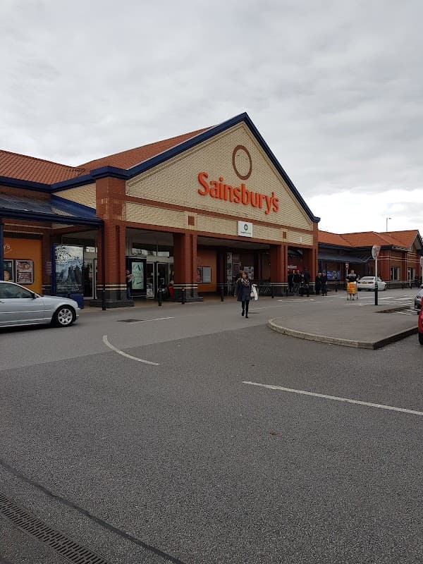 Sainsbury's storefront in Colton, Yorkshire, with shoppers and cars in the parking lot under a cloudy sky.