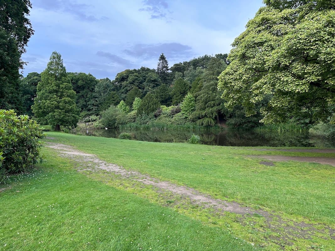 Lush green park with a path leading to a tranquil pond, surrounded by trees and shrubs under a cloudy sky.