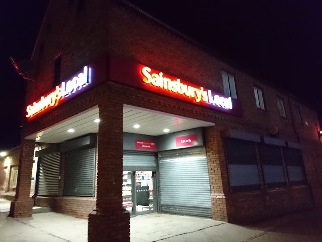 Sainsbury's Local storefront at night, featuring illuminated signage and closed shutters on windows.
