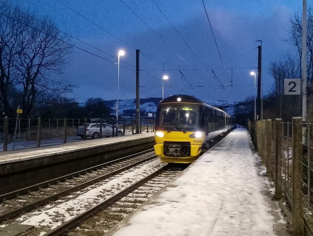 Train arriving at Cononley station, snowy platform, evening lights, and trees in the background.