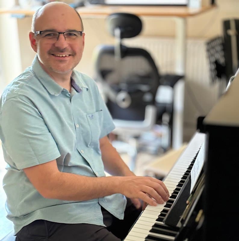 A man with glasses smiles while playing a piano in a music shop setting with chairs and music stands in the background.