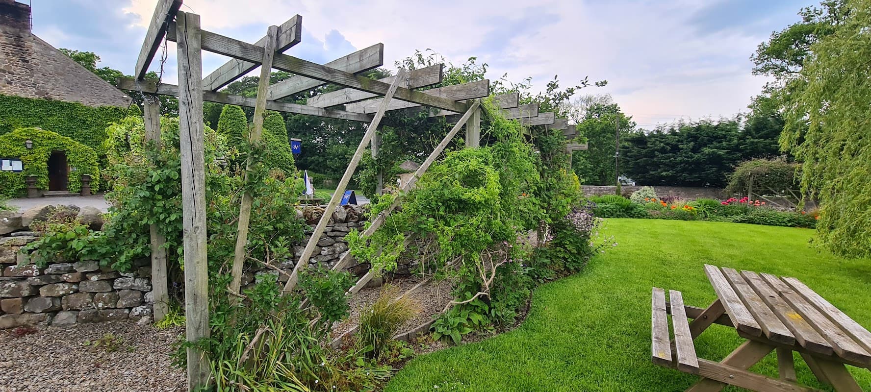 Wooden pergola covered in greenery, surrounded by a well-kept garden and stone wall at The Wyvill Arms.