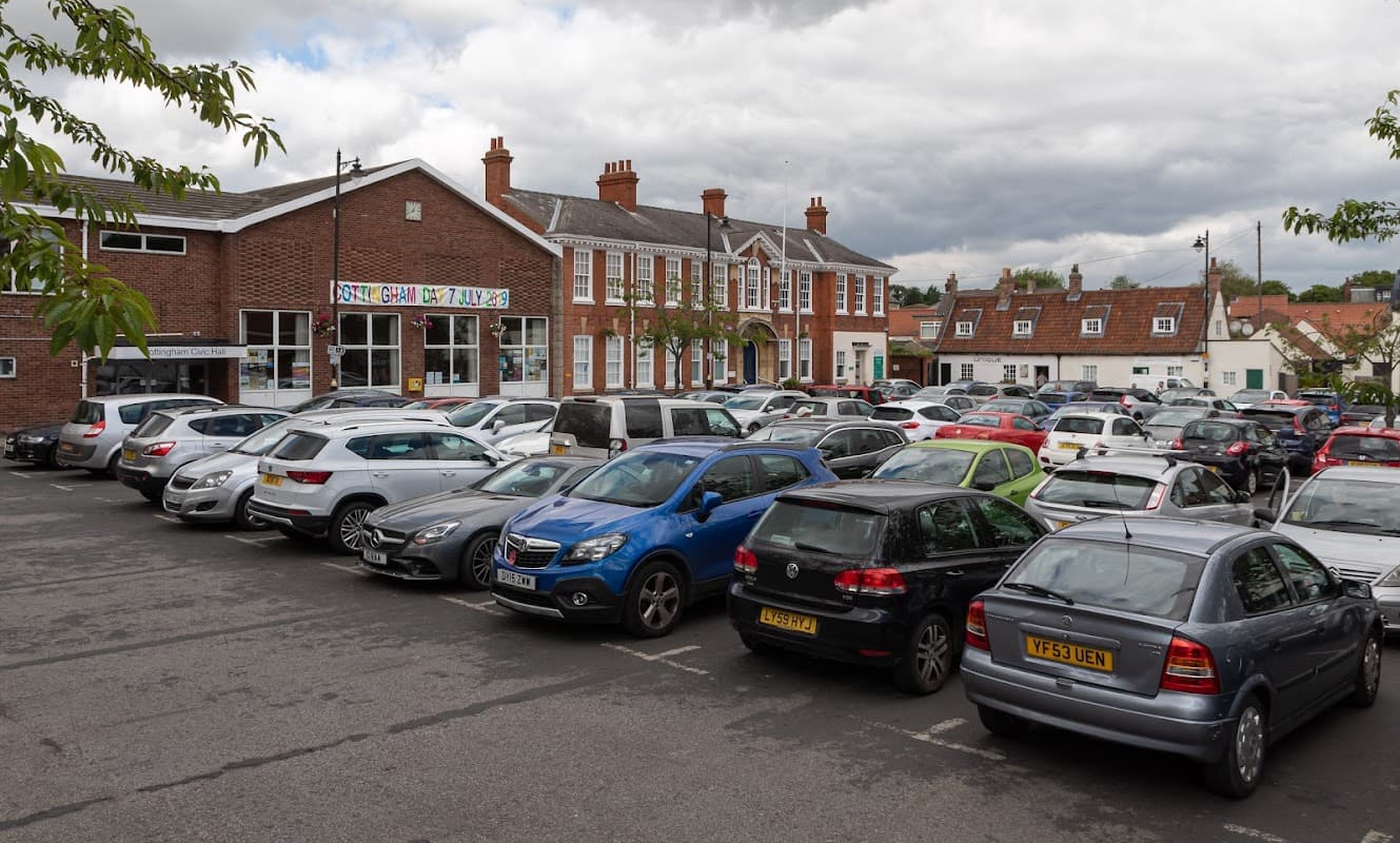 Busy Cottingham Green Car Park filled with parked cars, adjacent to a brick building and residential homes under a cloudy sky.