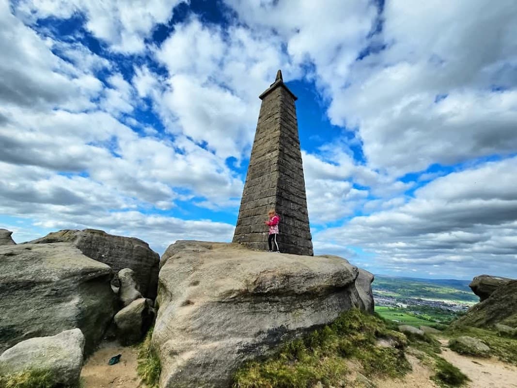 A tall stone monument stands on rocky terrain under a cloudy sky, with a person in pink near its base.