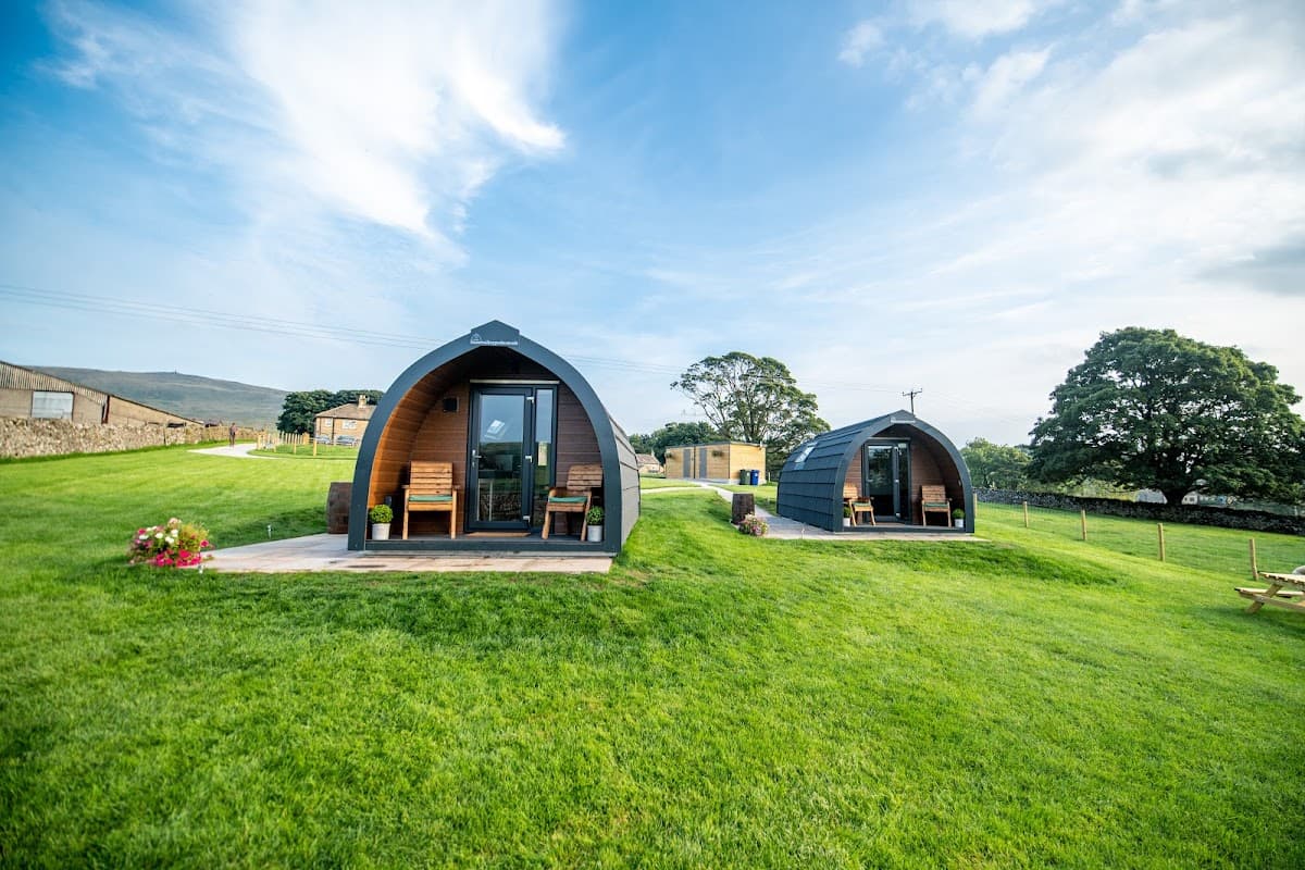 Two glamping pods with wooden decks sit on a grassy hill, surrounded by trees and scenic Yorkshire Dales landscape.