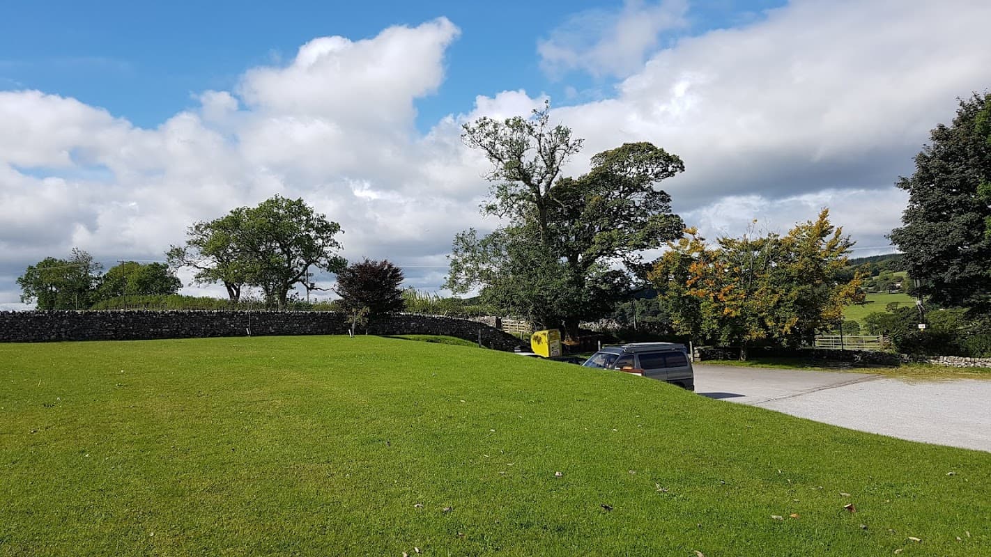 Cracoe Village Hall surrounded by green grass, trees, and a stone wall under a partly cloudy sky.