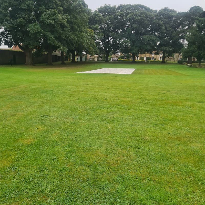 Green cricket field with a white pitch cover and large trees in the background.