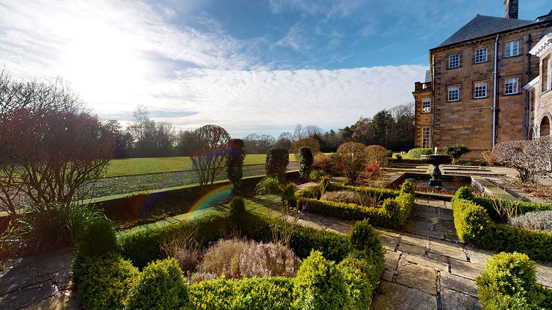 Elegant gardens with neatly trimmed hedges and stone pathways, framed by Crathorne Hall's historic architecture and a sunny sky.