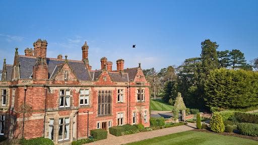 Victorian-style red brick mansion with ornate architecture, surrounded by lush greenery and a clear blue sky.
