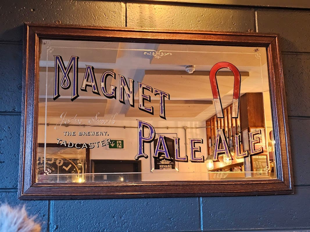 Framed sign featuring "Magnet Pale Ale" with a red magnet illustration, set against a rustic pub interior.