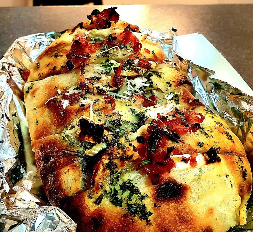 Golden, crusty bread topped with herbs, cheese, and crispy bits, served in a foil tray at New Yorkshire Food Co.