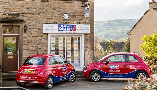Two red cars parked outside a stone building with a sign for "Wilman & Lodge" and property listings in the windows.