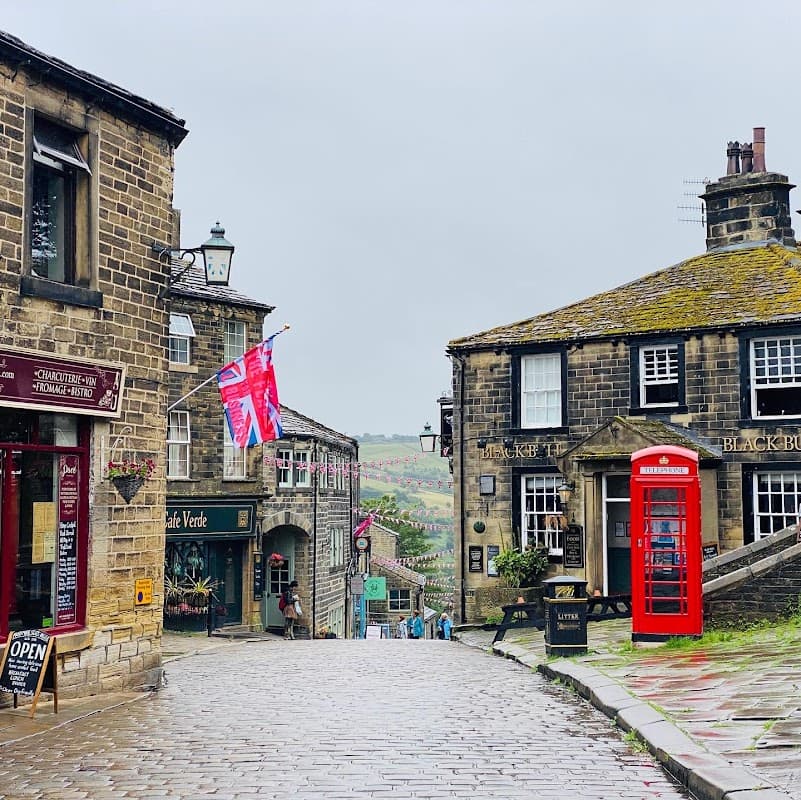 Quaint cobblestone street with shops, a red telephone box, and flags, surrounded by green hills under a cloudy sky.