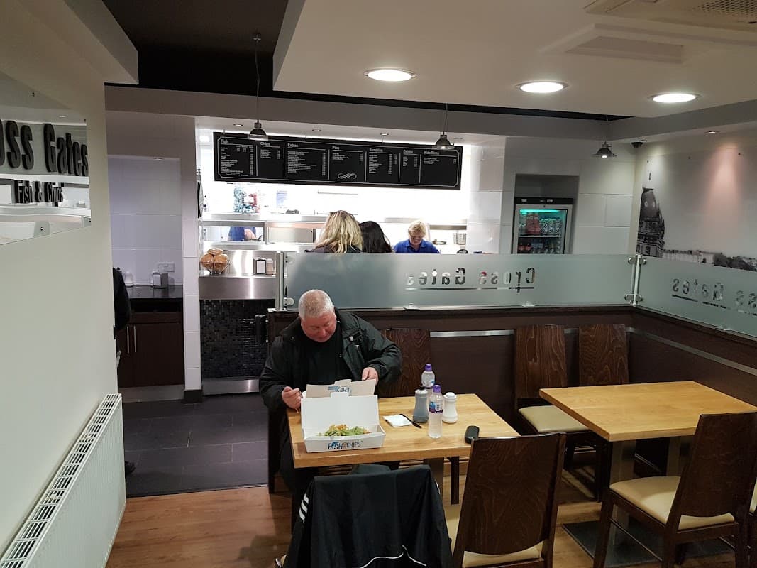 A man sits at a wooden table enjoying fish and chips in a modern restaurant with a menu board and staff behind the counter.