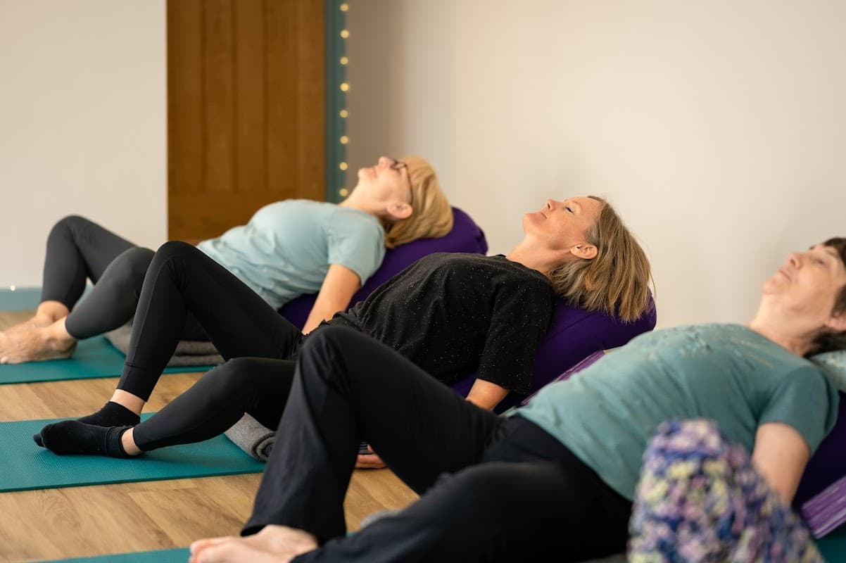 Three women lying on yoga mats, using bolsters for support, in a serene studio setting.