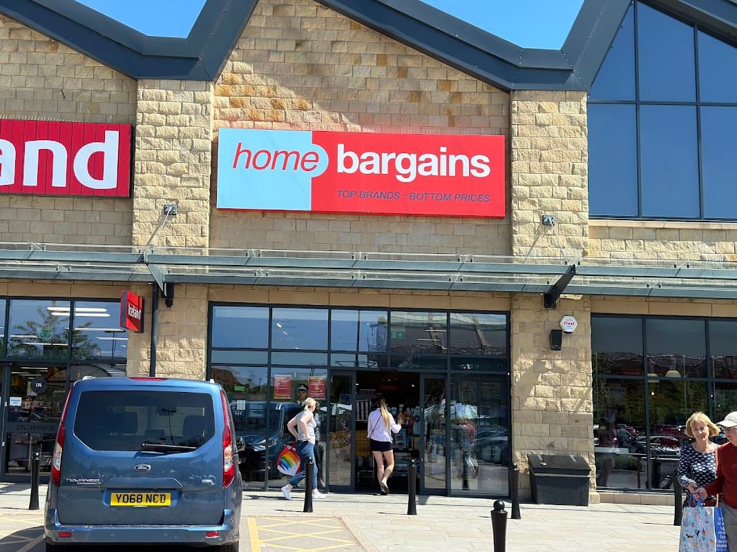 Storefront of Home Bargains with a large red sign, shoppers entering, and a blue vehicle parked outside.