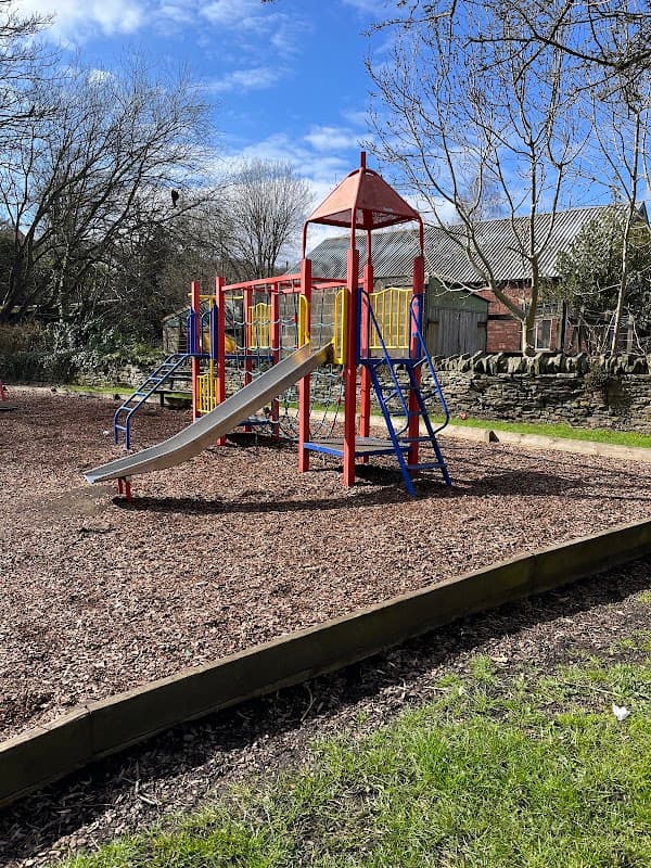 Colorful playground equipment with a slide, surrounded by wood chips and trees in Denby Dale, Yorkshire.