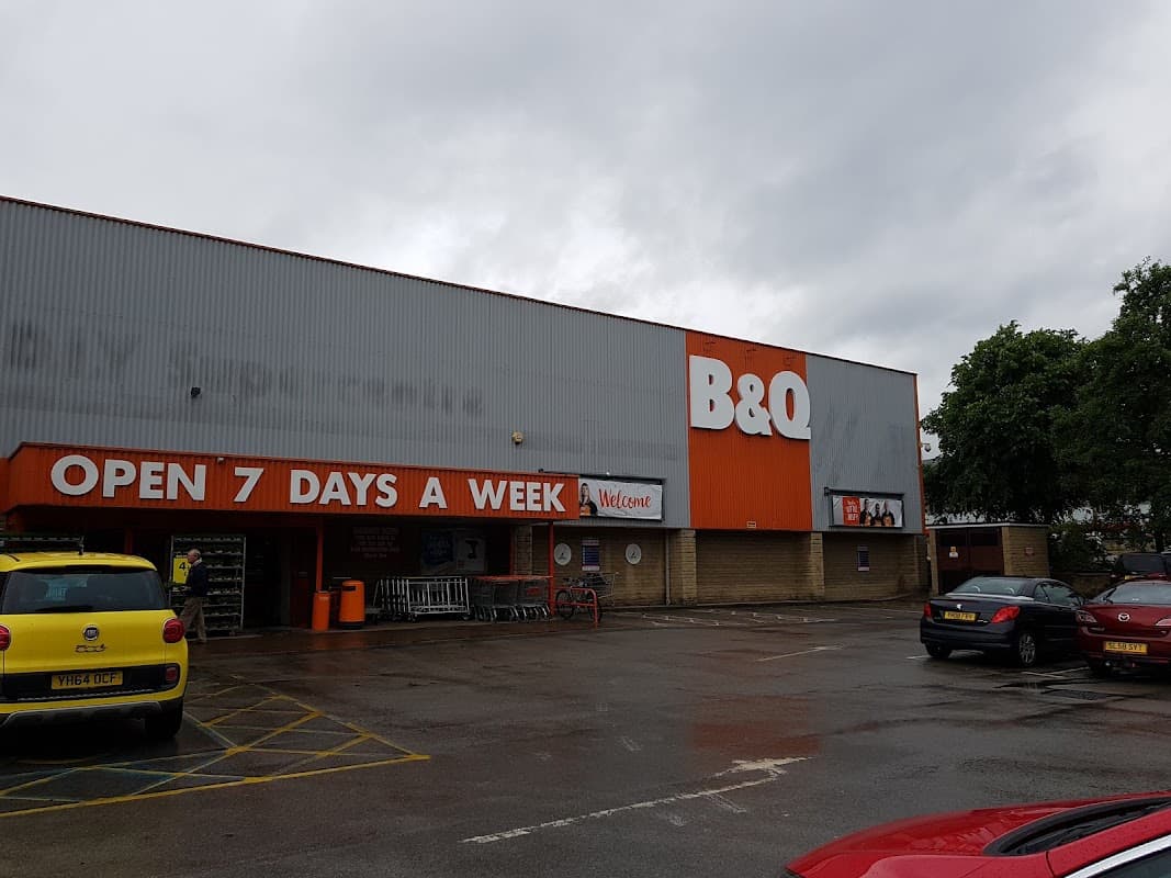 B&Q store exterior in Dewsbury with a large orange sign, parking area, and cloudy sky. Open 7 days a week banner visible.