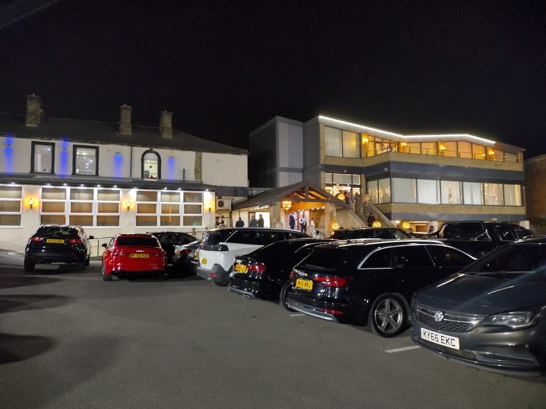 Car park at night with several parked cars, a modern building with bright lights, and a traditional structure nearby.