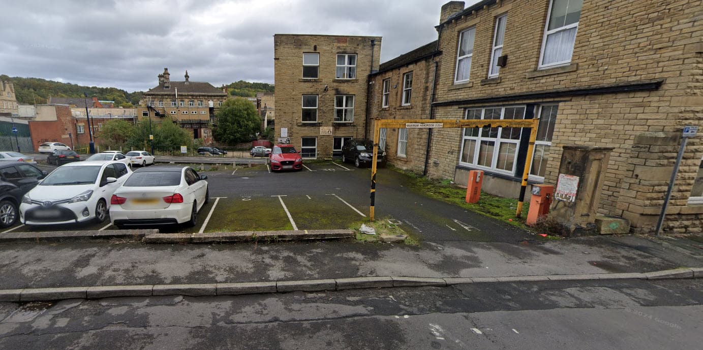 Car park with several vehicles, stone buildings in the background, and a cloudy sky in Dewsbury, Yorkshire.