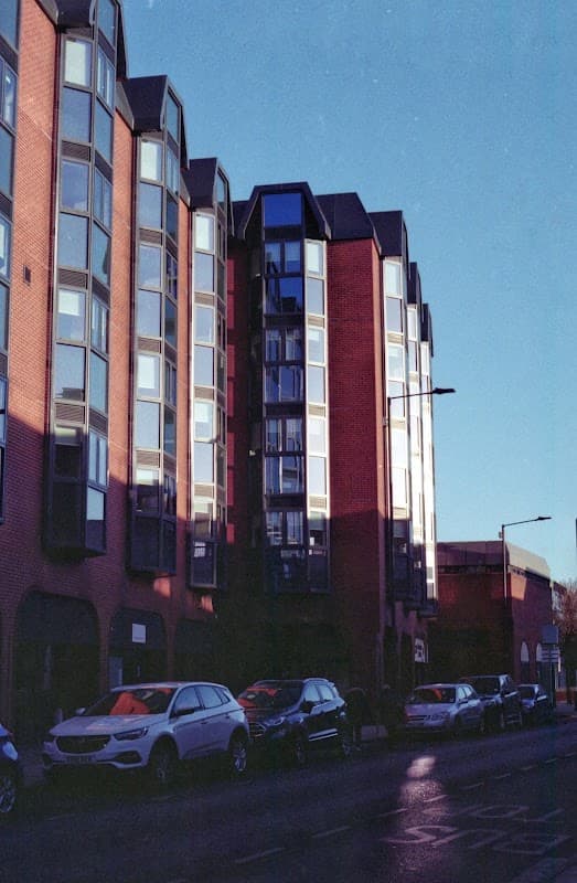 Modern brick building with large windows, reflecting sunlight, alongside parked cars on a city street.