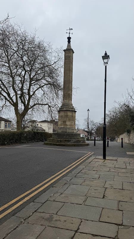 Stone column with a weather vane, surrounded by trees and lampposts on a cobblestone street in Doncaster.
