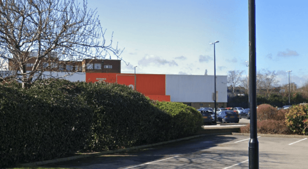 Car park with parked cars, hedges, and a building featuring orange and white panels in Dudley Hill, Yorkshire.