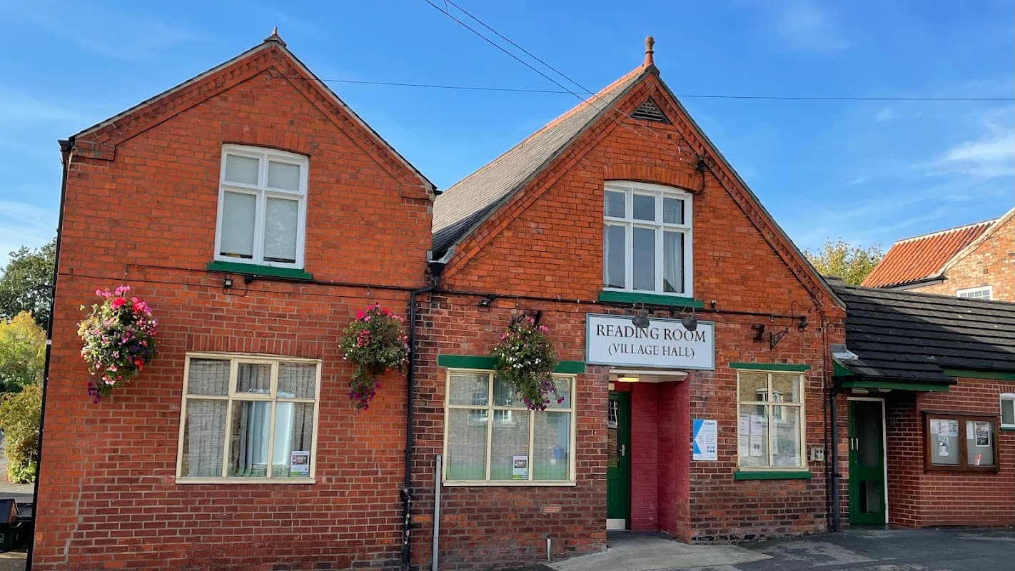 Red brick building with a gabled roof, green accents, and flower baskets, labeled "Reading Room (Village Hall)."