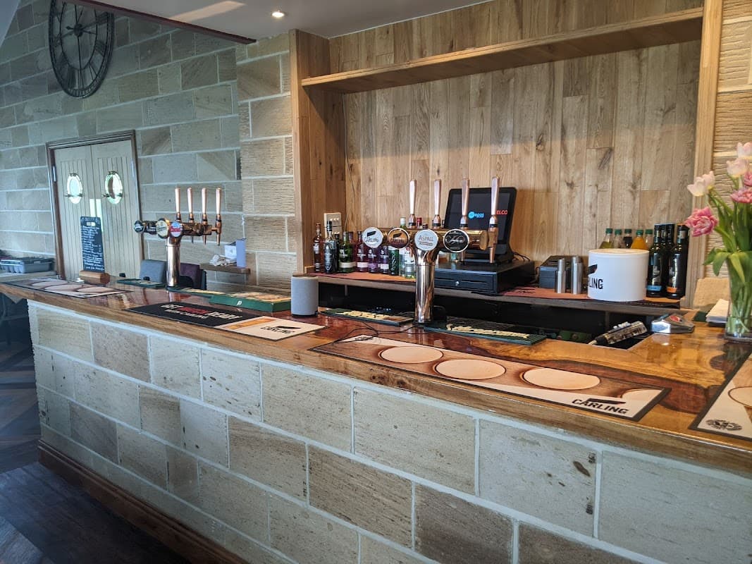 Wooden bar with beer taps, bottles, and glasses; stone wall and floral decorations in a cozy pub setting.