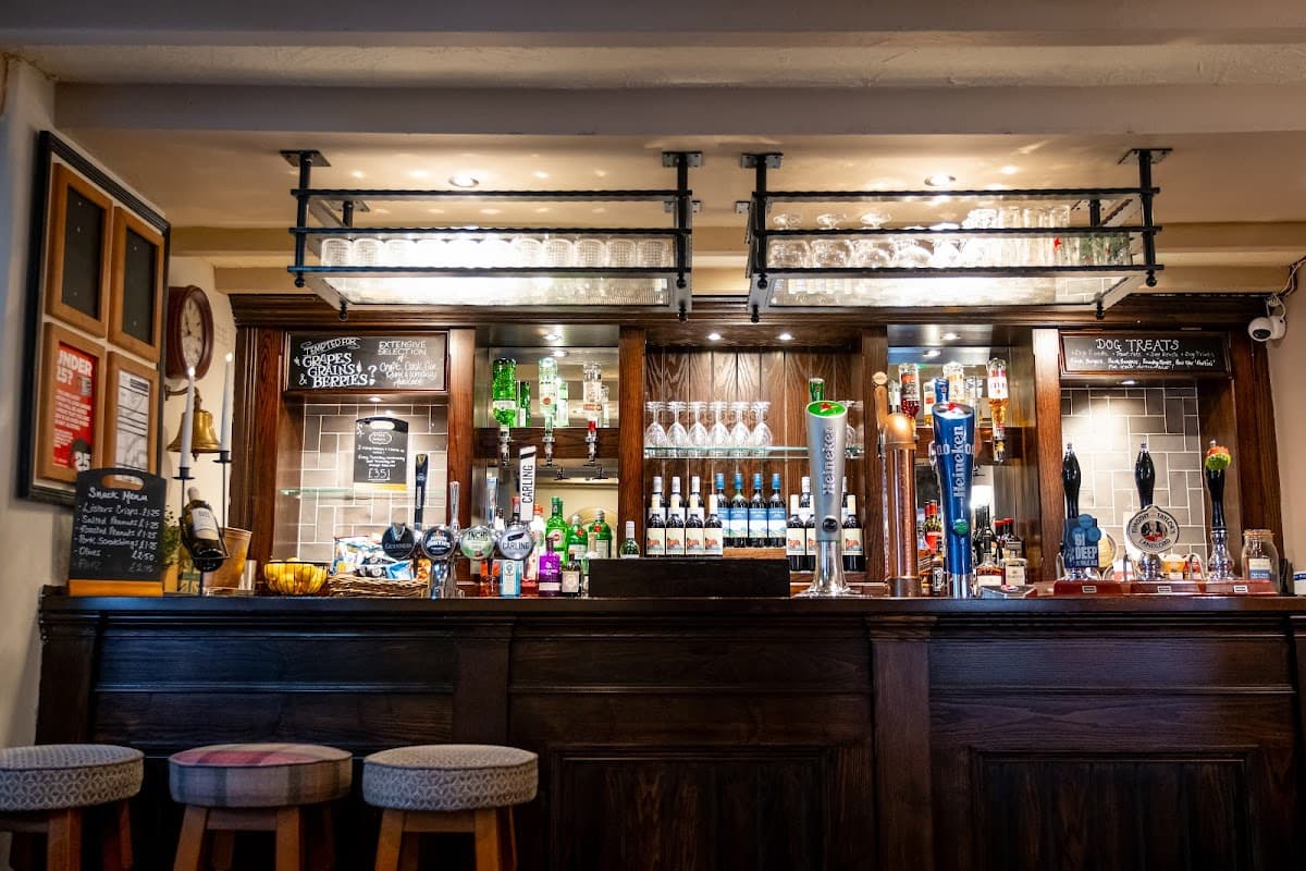 Wooden bar with various bottles, glasses, and taps; chalkboard menus and stools in a cozy pub setting.