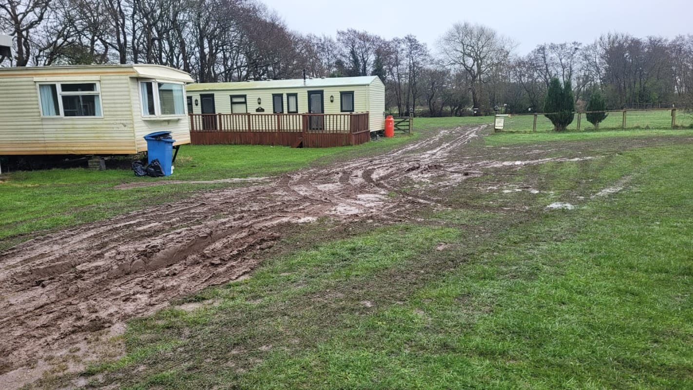 Muddy path leading to static caravans, with grassy areas and trees in the background at Riverside Camp Site.