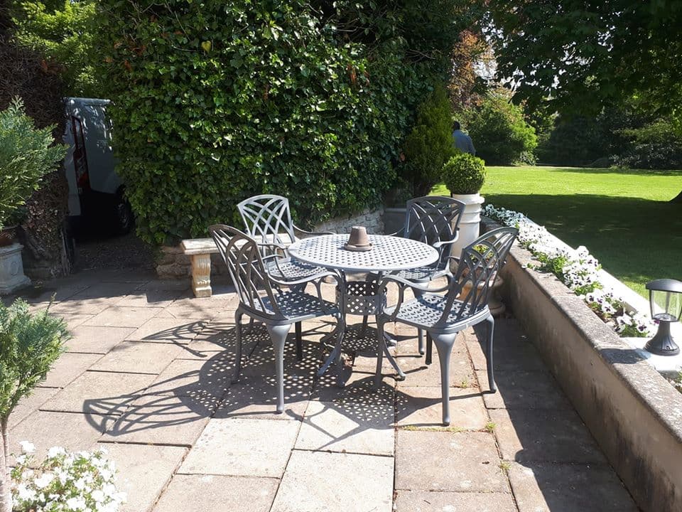 Outdoor dining area with a round table and four chairs, surrounded by greenery and a stone pathway.