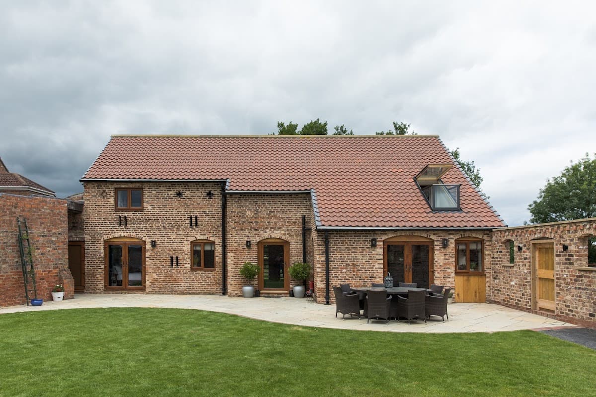 Charming brick barn hotel with a red tiled roof, outdoor seating area, and lush green lawn under a cloudy sky.