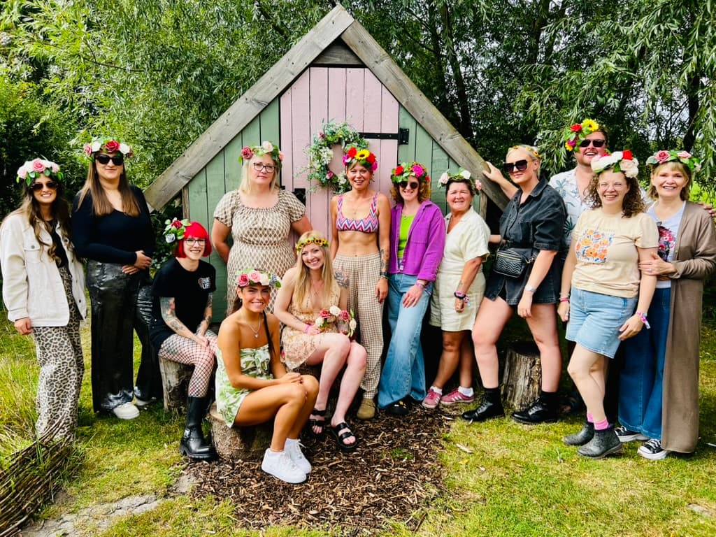 Group of women in floral crowns and stylish outfits posing cheerfully outside a colorful wooden cabin in a garden setting.